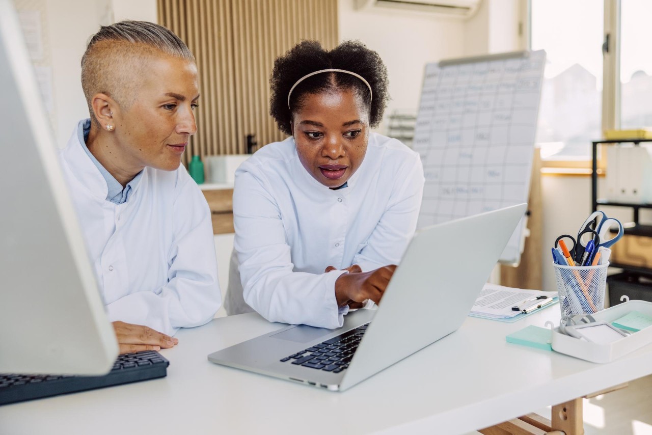 Front view of two female HCPs leaning on a standing desk, one is pointing to the monitor and the other is looking at the monitor; office supplies are on the desk next to another desktop and keyboard; blurry in the background is an easel with a hand-drawn chart.